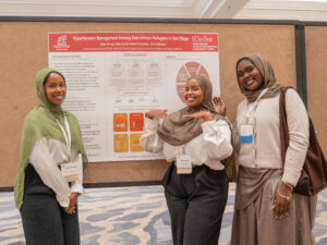 Three smiling researchers pose beside their poster on hypertension management among East African refugees in San Diego at the 2026 International Refugee and Migration Health Conference