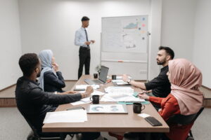 Five colleagues sit around a conference table covered with printed charts and notebooks, listening to a coworker presenting at a whiteboard.