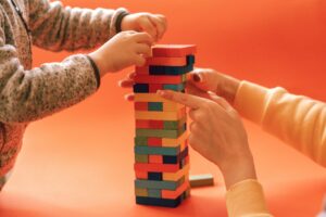alt="A child and an adult stacking colorful wooden blocks into a tall tower together against an orange background"