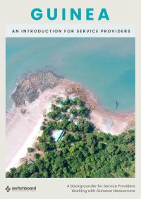 Cover of a Switchboard resource titled 'Guinea: An Introduction for Service Providers — A Backgrounder for Service Providers Working with Guinean Newcomers.' The cover shows an aerial photograph of a small forested peninsula in Conakry, Guinea, with green trees, pale sand, and turquoise ocean water. Two long boats are moored along the shore.