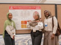 Three smiling researchers pose beside their poster on hypertension management among East African refugees in San Diego at the 2026 International Refugee and Migration Health Conference