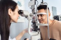 An optometrist examines a young man's eyes using a slit lamp.