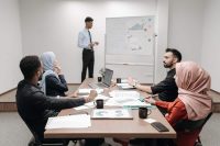 Five colleagues sit around a conference table covered with printed charts and notebooks, listening to a coworker presenting at a whiteboard.